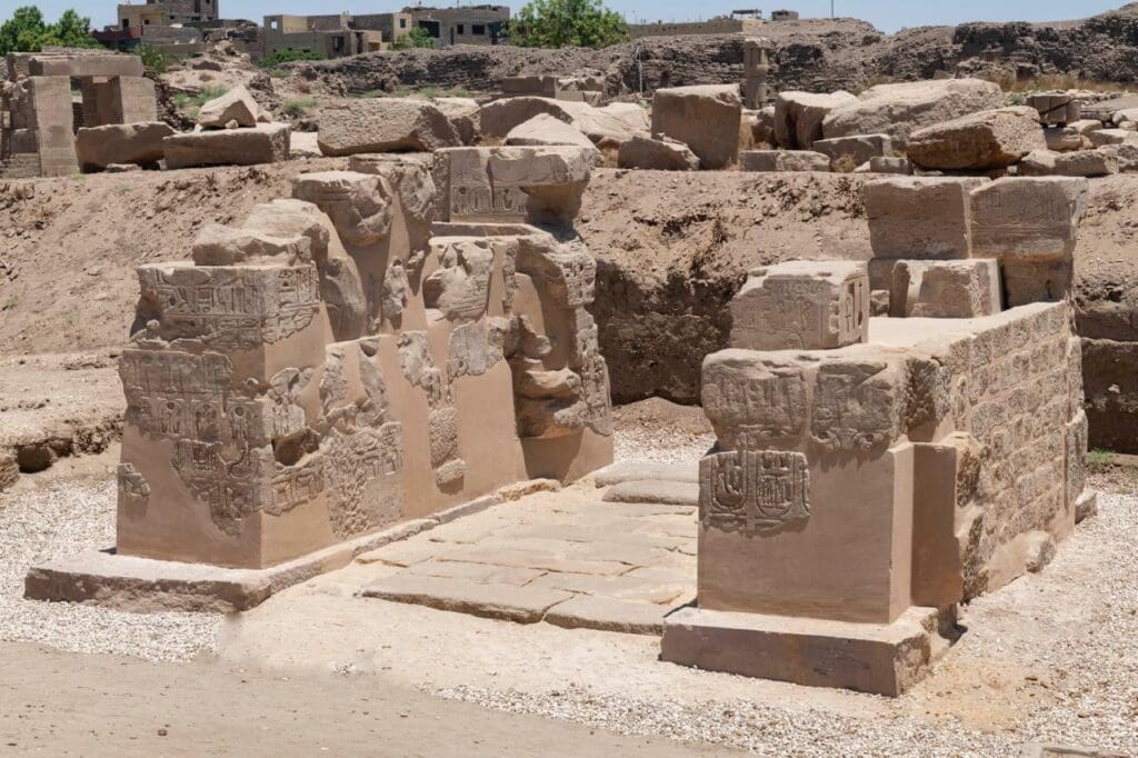Remains on ancient stone walls, intricately carved, a few feet tall, with a stone floor and several large hewn rocks and other modern buildings in the background. Photo courtesy the Egyptian Ministry of Tourism and Antiquities