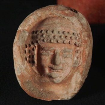 Circular mold with feminine face inside with three rows of curls over the forehead and covering the ears. Face has naturalistic features. Courtesy Israel Antiquities Authority.