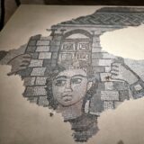 mosaic of samson with black curly hair and no beard with brickes behind him and gates above his head, arms raised. Courtesy Israel Antiquities Authority